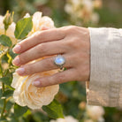 [KI] Silberring mit Mondstein an der Hand zwischen Blumen