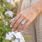 [KI] Silberring mit Mondstein an der Hand zwischen Blumen